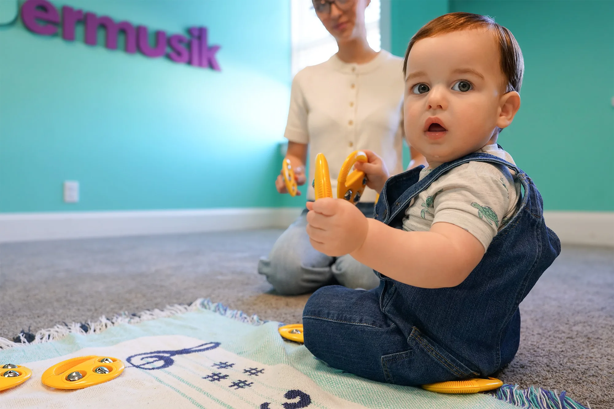Baby With Yellow Jingle Bells A baby in denim overalls holds up yellow jingle bells on a music-note blanket as a grown-up plays along