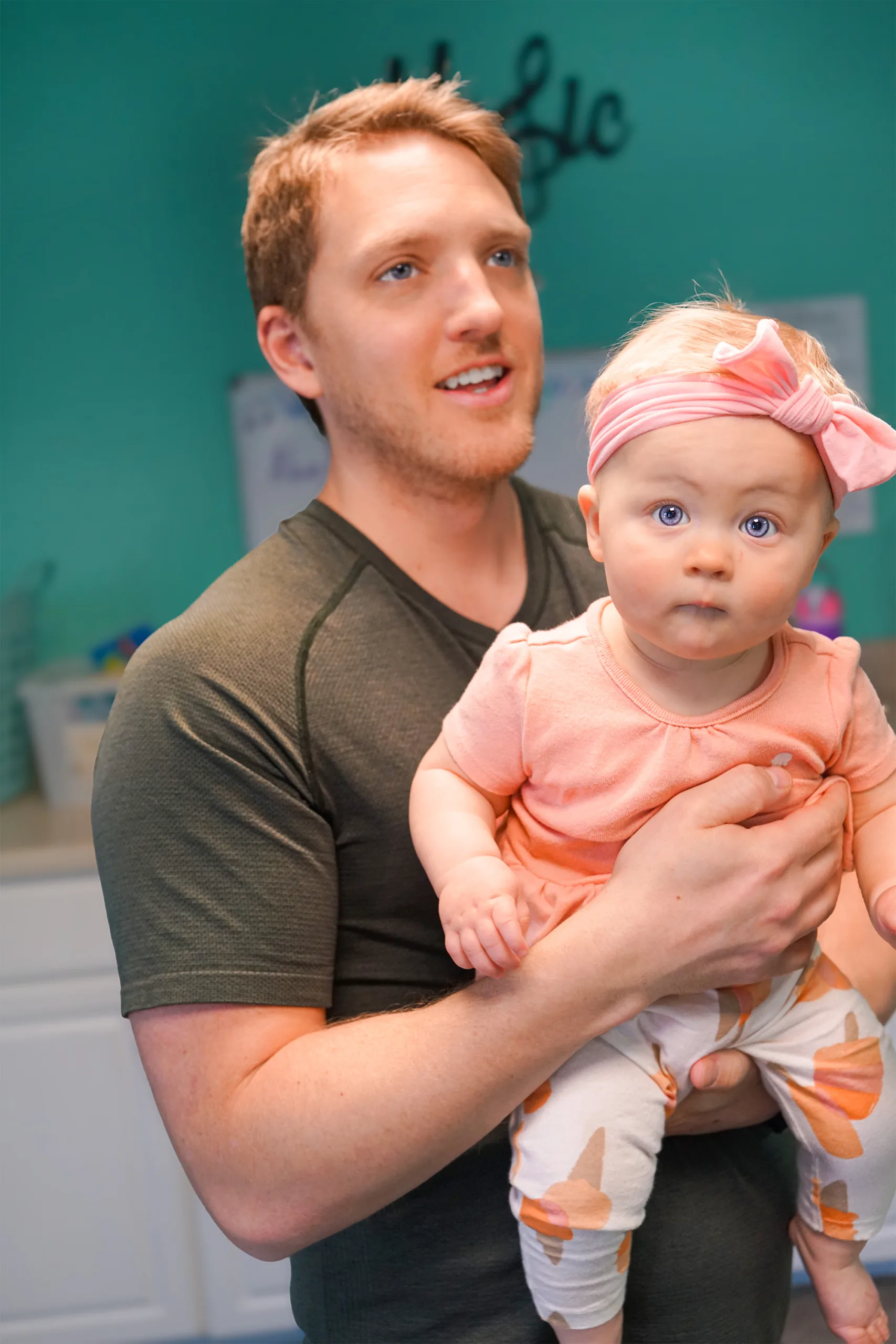 Man Holding Baby In Pink Headband A smiling man holds a wide-eyed baby in a pink bow headband during class