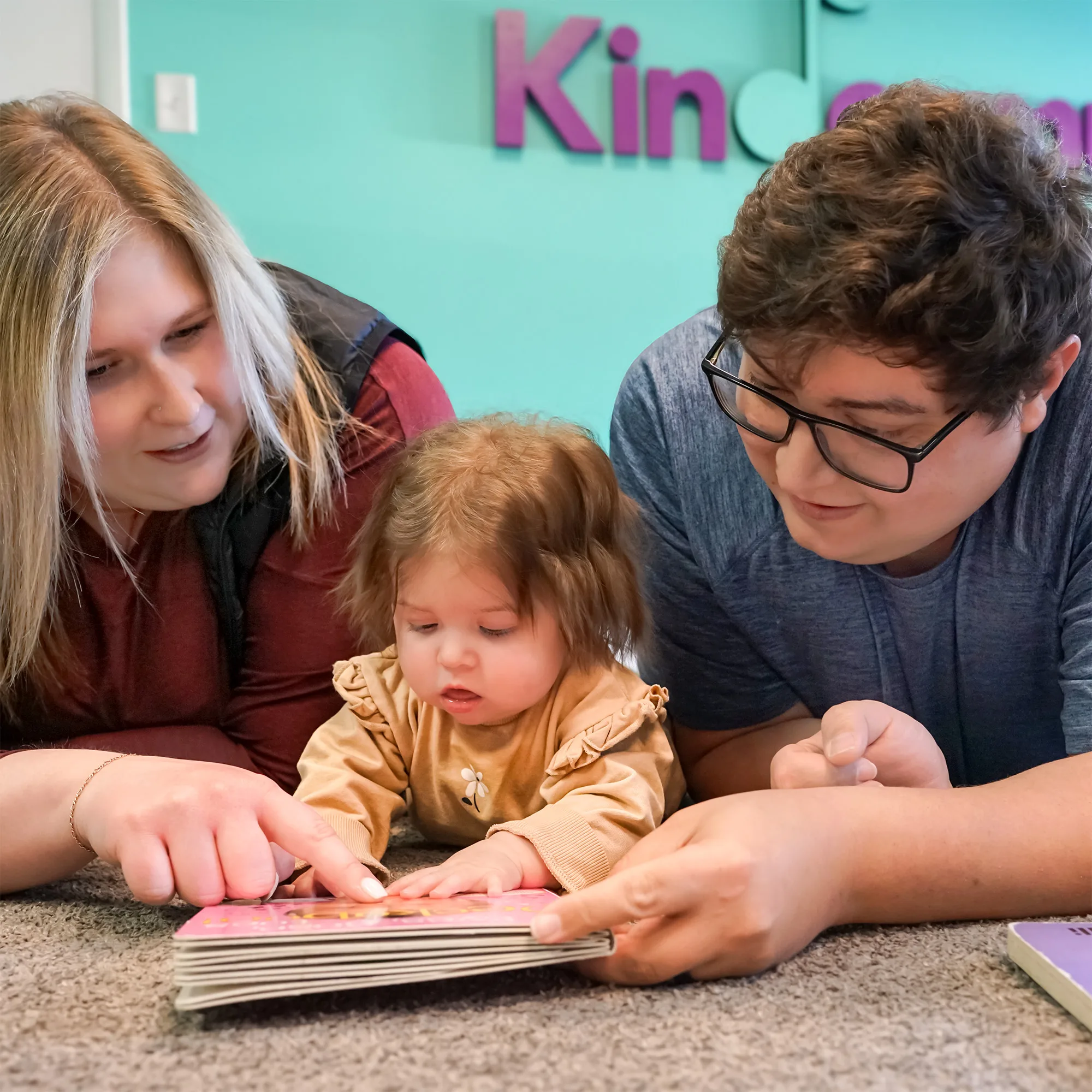 Baby And Grown-Ups Reading Board Book Two grown-ups lie on the carpet sharing a board book with a baby in a Kindermusik class
