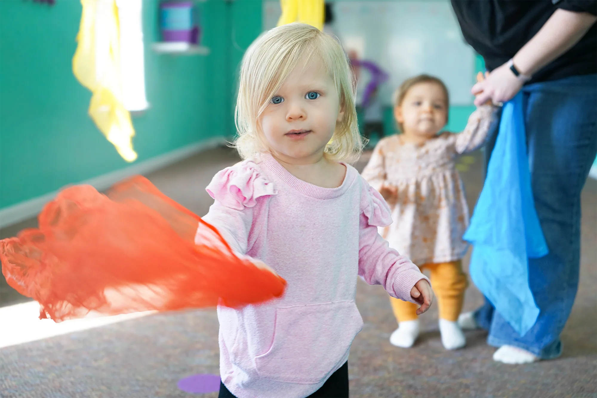 Toddler Waving Orange Scarf A toddler in pink waves a sheer orange scarf while another toddler walks behind holding a grown-up's hand