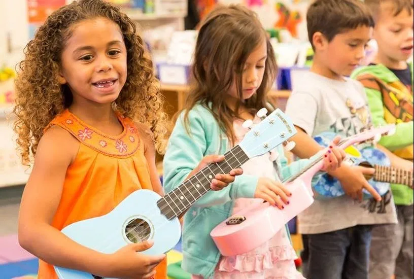 Children Playing Ukuleles In A Row Four young children stand in a row playing ukuleles, a smiling girl in orange at the front