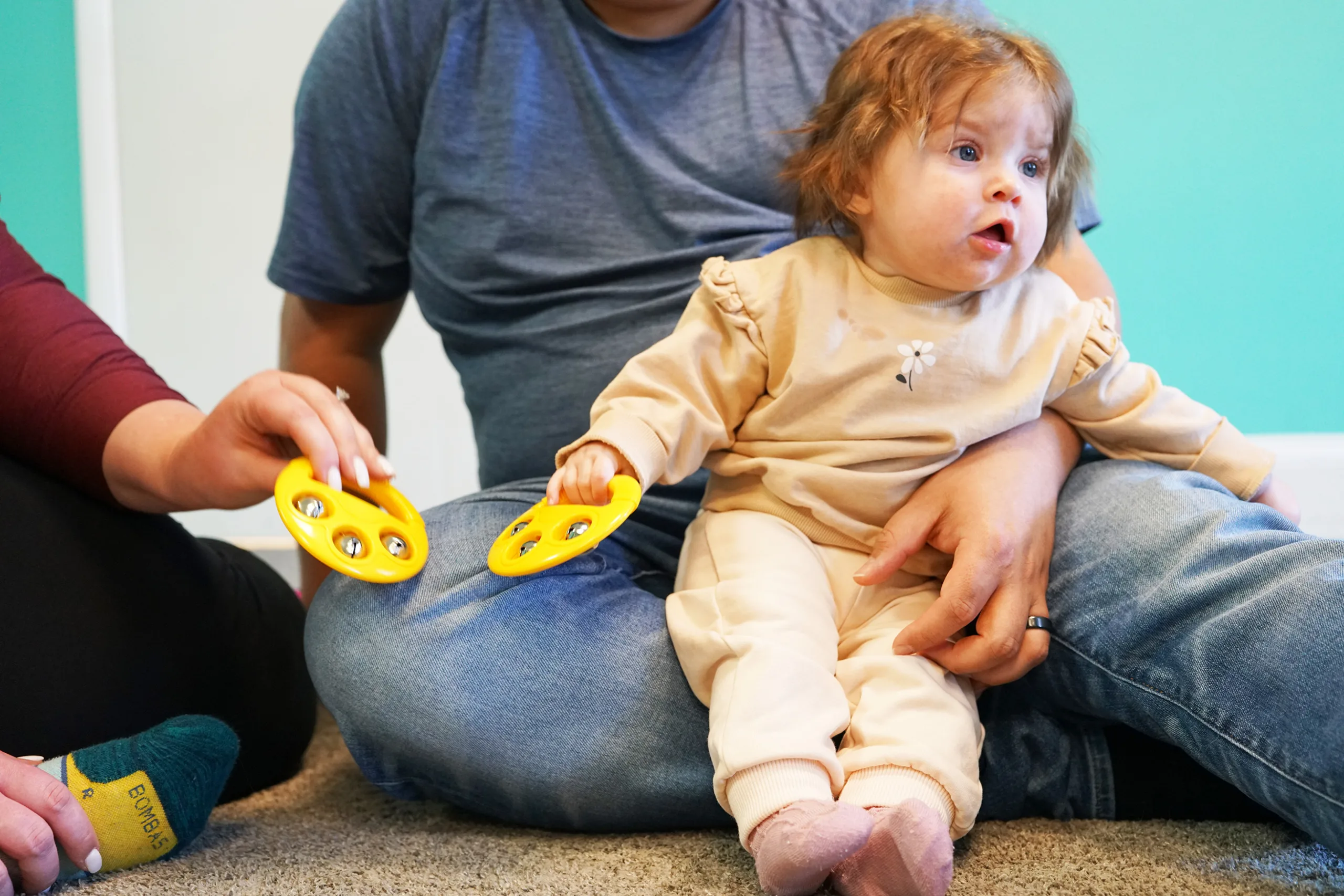 A baby on a grown-up's lap holds a yellow jingle bell as another adult shakes a matching one