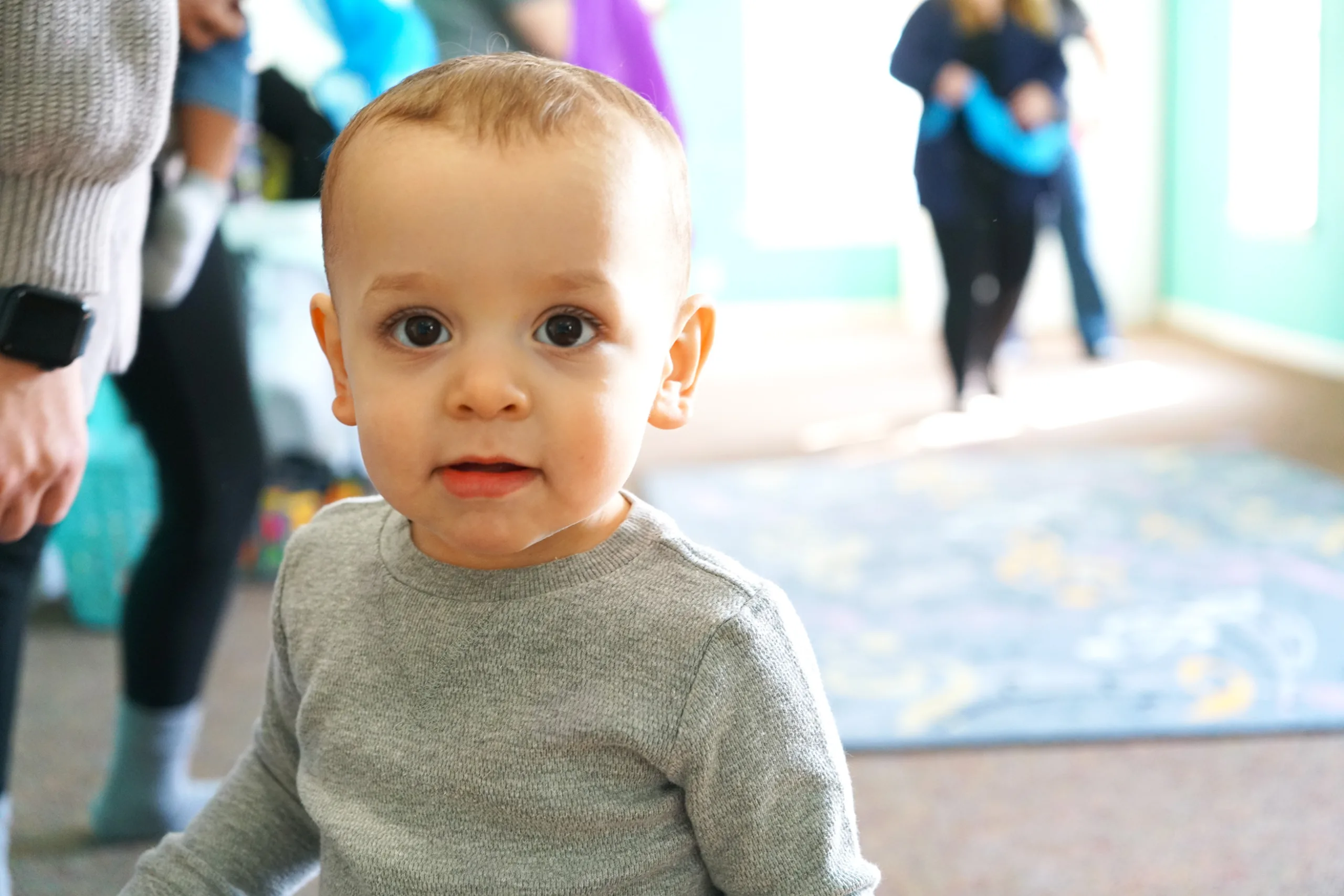 Baby In Gray Sweater Close-Up A wide-eyed baby in a gray sweater looks into the camera as grown-ups move around the room behind