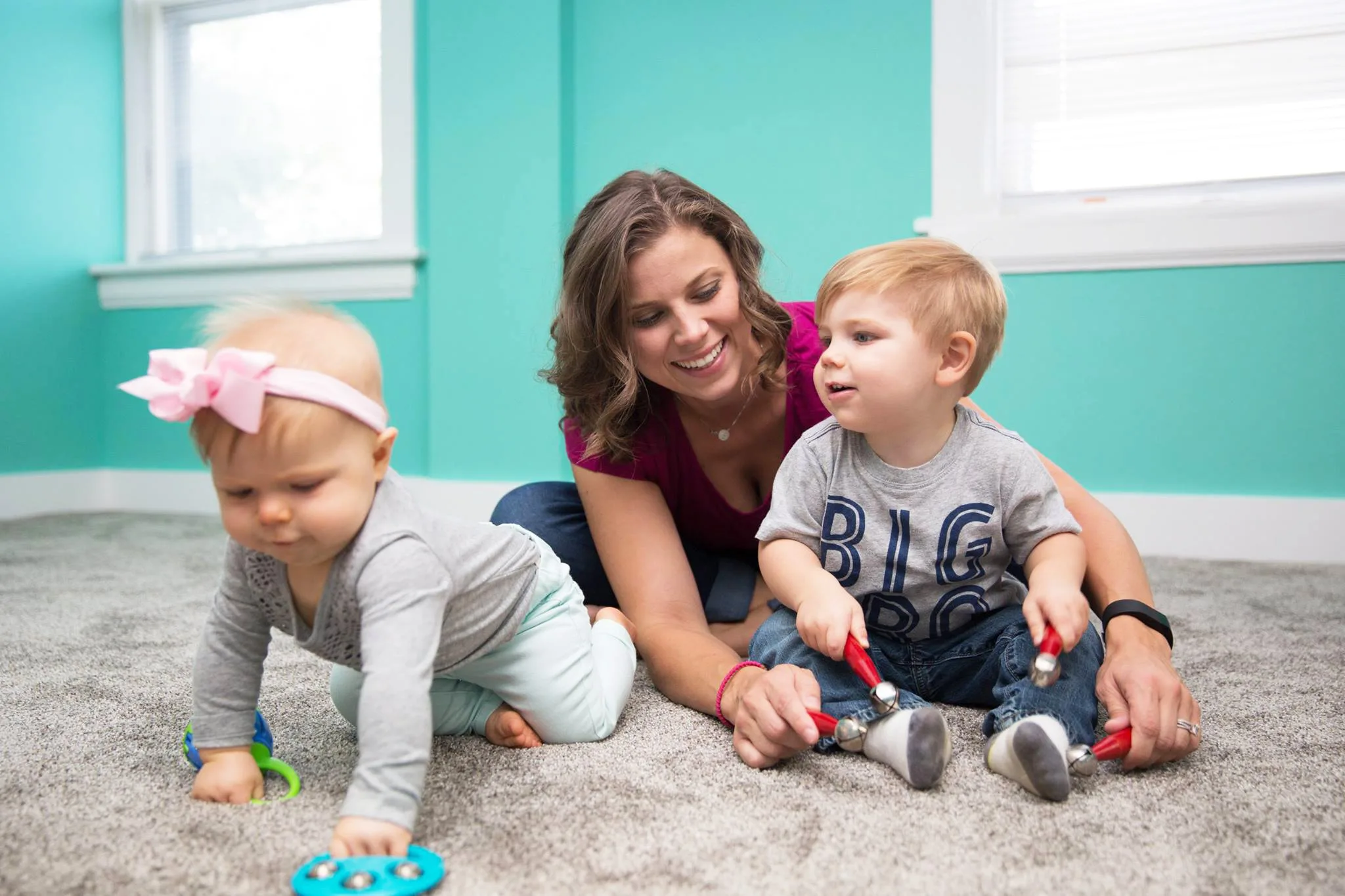 A smiling woman on the carpet with a toddler shaking red jingle-bell sticks and a crawling baby in a pink bow