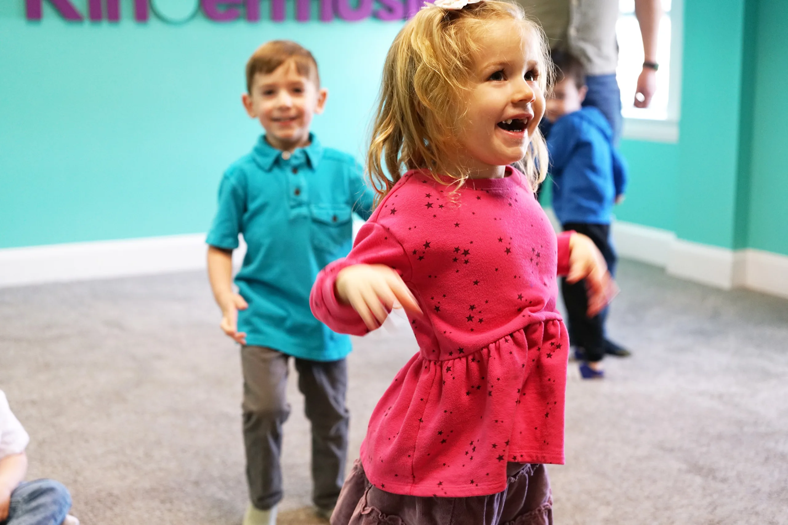 A laughing girl in a pink star-print top dances across the room, a smiling boy following behind