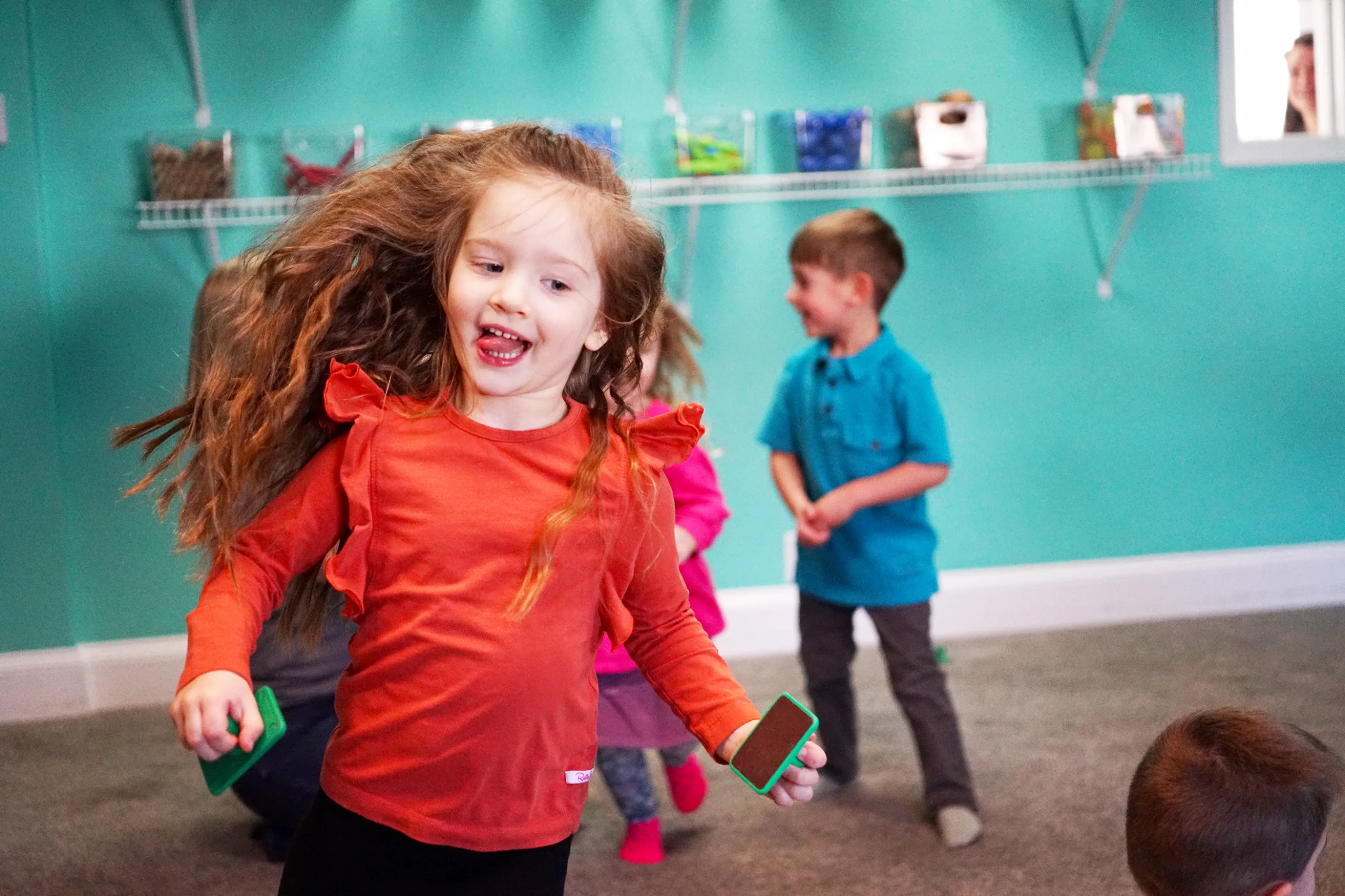 Girl Spinning With Hand Clappers A laughing girl spins with her hair flying, a green hand clapper in each hand, as classmates dance behind her