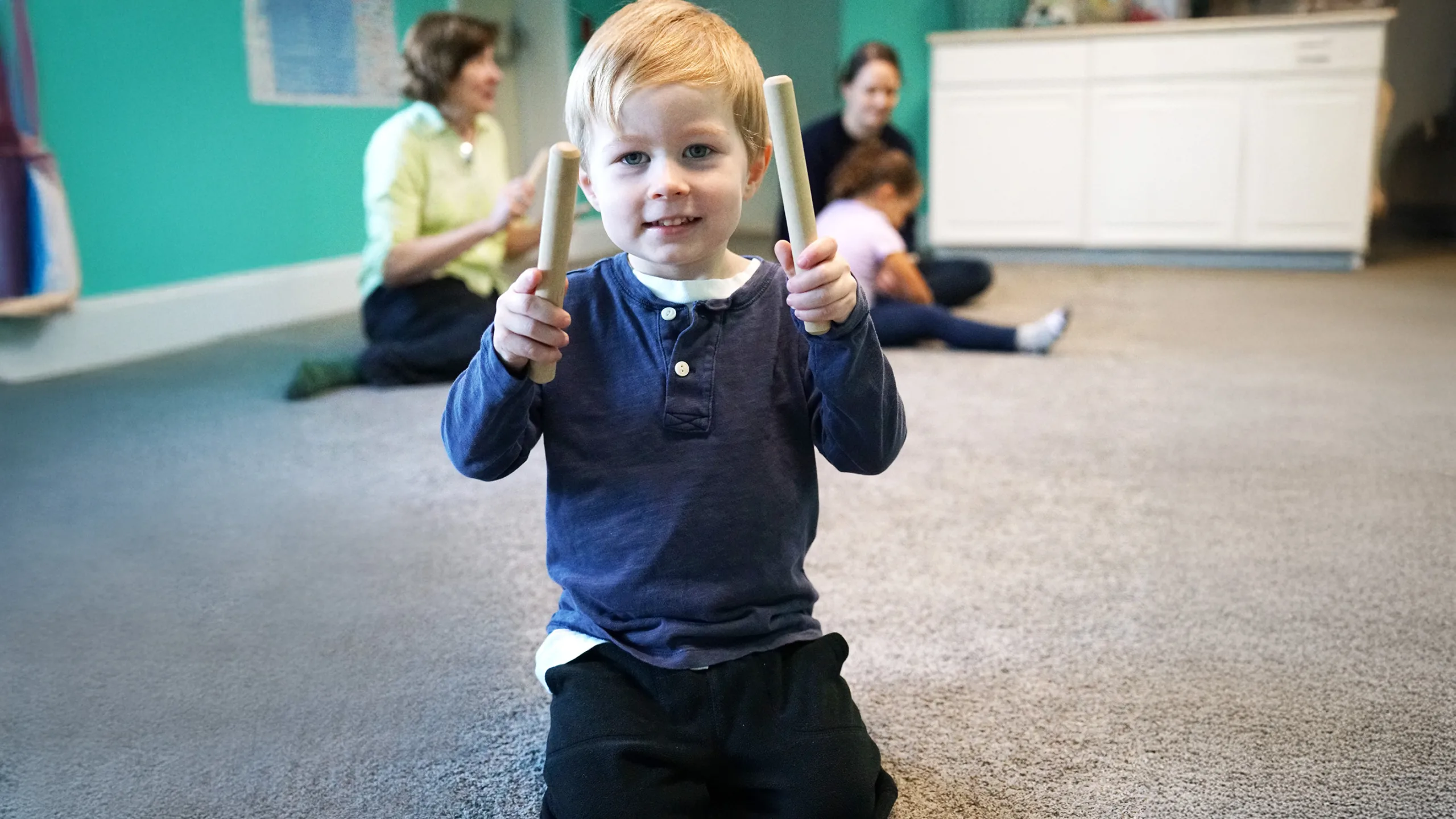 A smiling little boy kneels on the carpet holding up a wooden rhythm stick in each hand