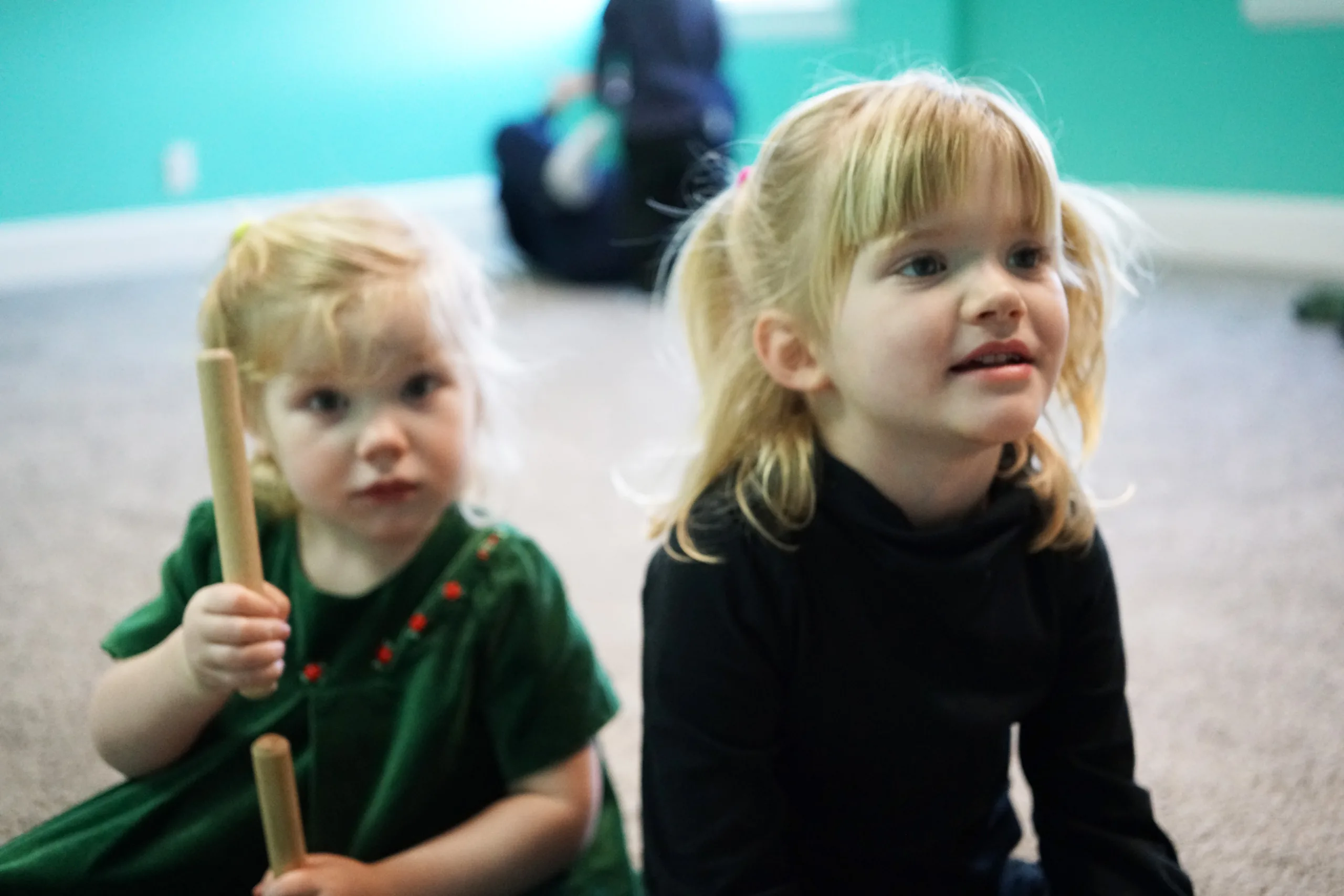 Two Girls With Rhythm Sticks Two little girls sit on the carpet looking ahead, the younger one holding a pair of wooden rhythm sticks