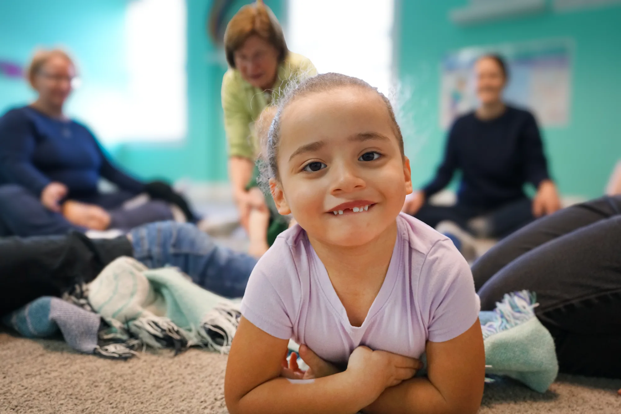 Grinning Girl Lying On Carpet A grinning girl lies on her tummy with her arms folded under her chin, grown-ups seated in a circle behind her