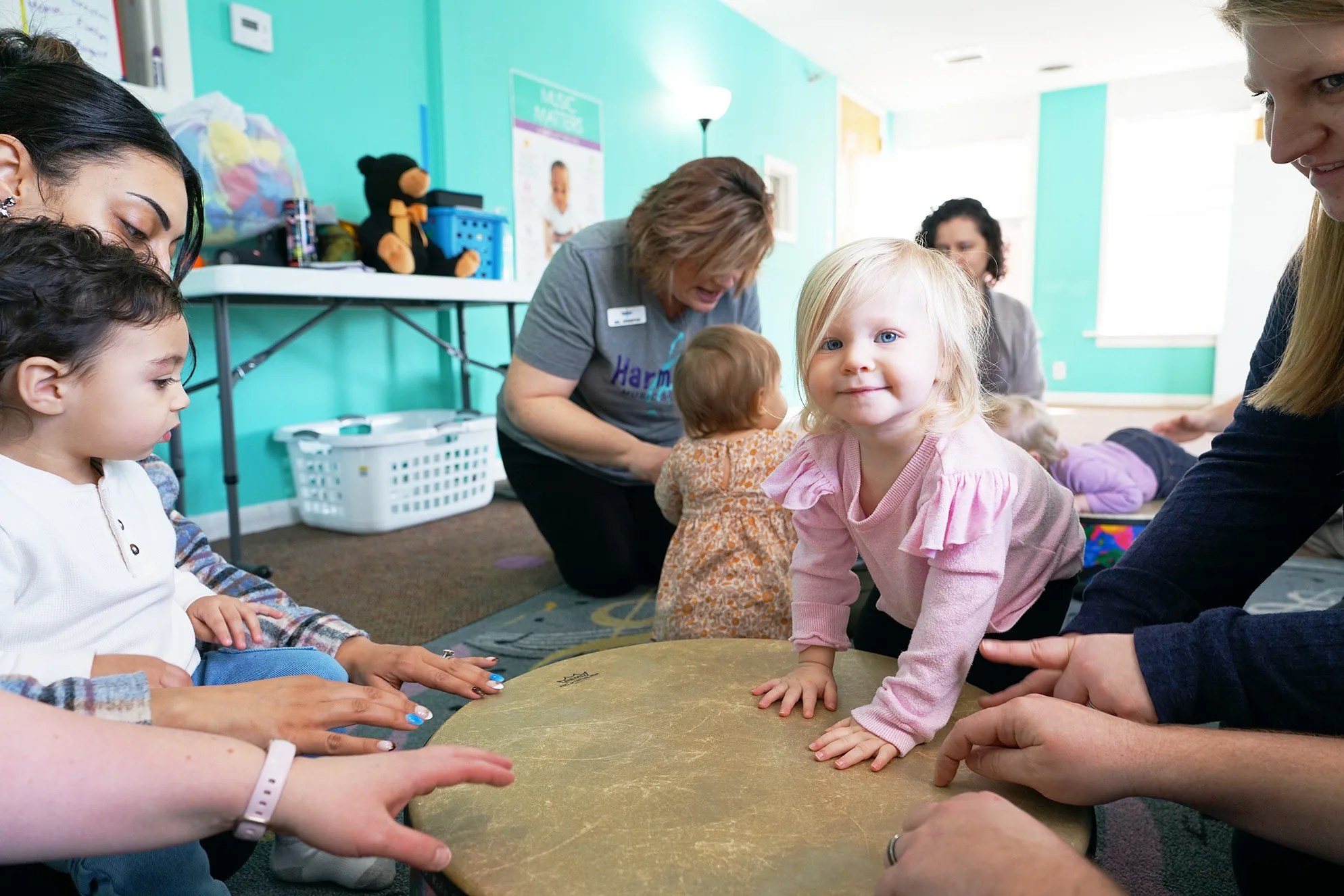 Toddlers and grown-ups gather around a large floor drum, a smiling girl in pink resting her hands on it
