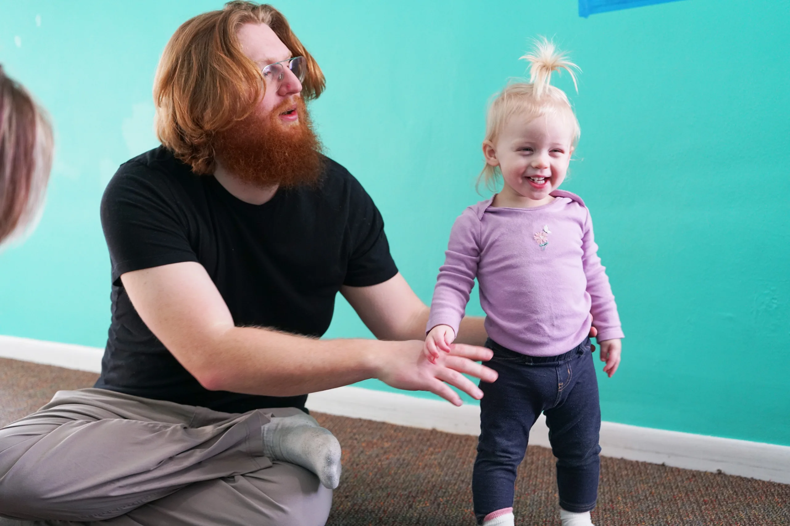 Laughing Toddler With Bearded Man A laughing toddler in lavender stands beside a bearded man seated on the floor, his hand held out to her