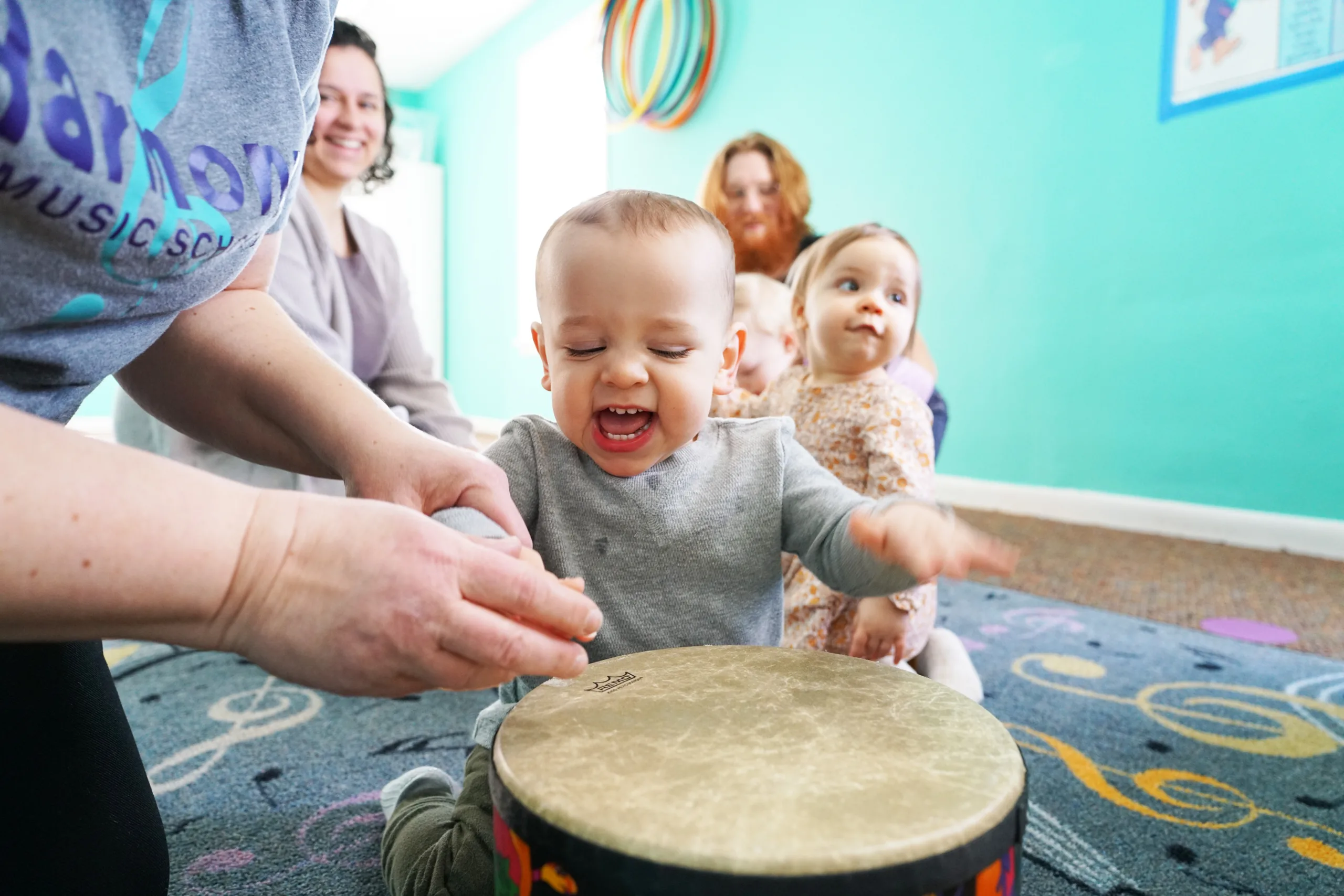 Laughing Baby Playing Hand Drum A laughing baby pats a hand drum on a music-note rug beside an adult in a Harmony Music School T-shirt