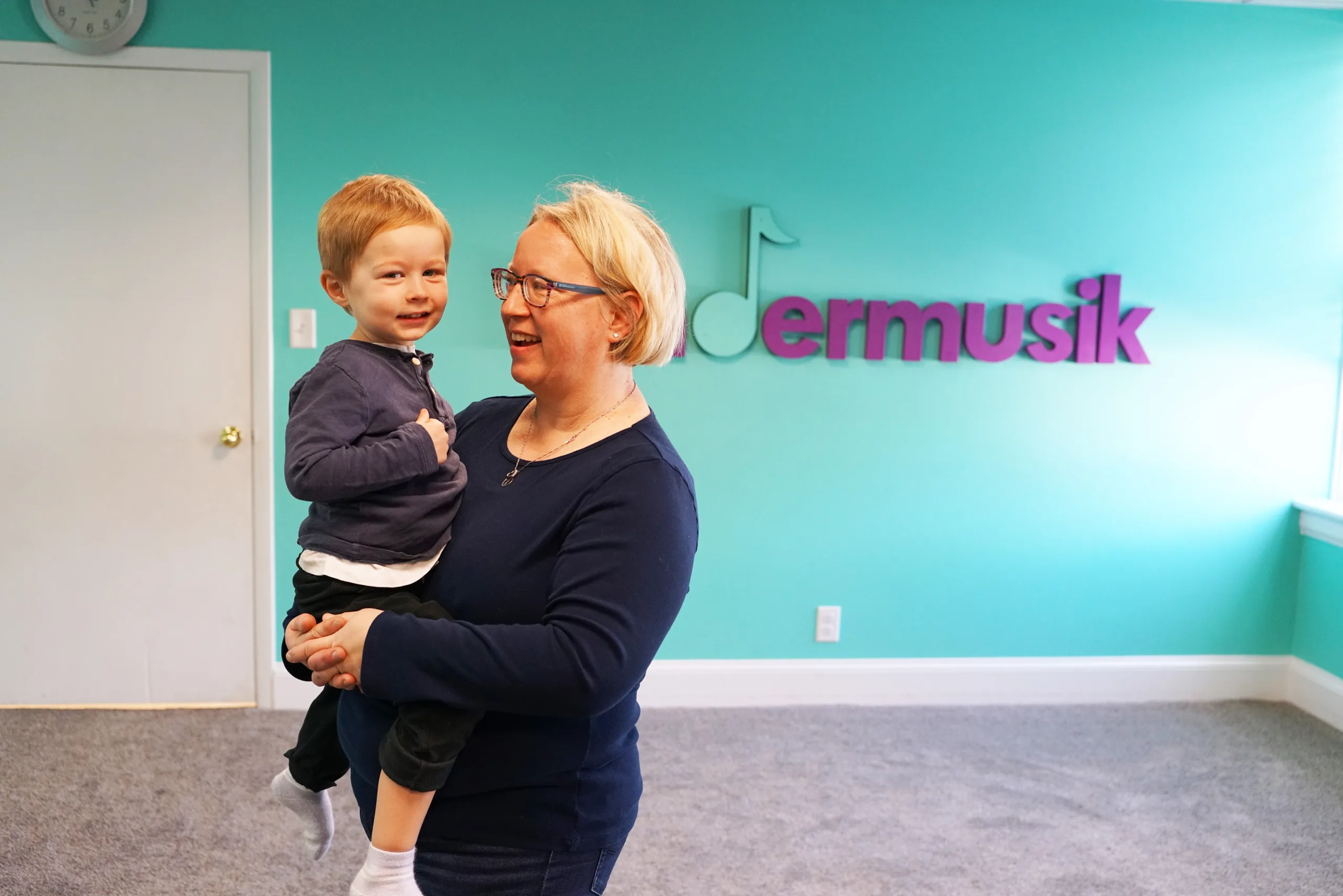 Woman Holding Toddler By Kindermusik Sign A smiling woman holds a red-haired toddler on her hip in front of the Kindermusik sign