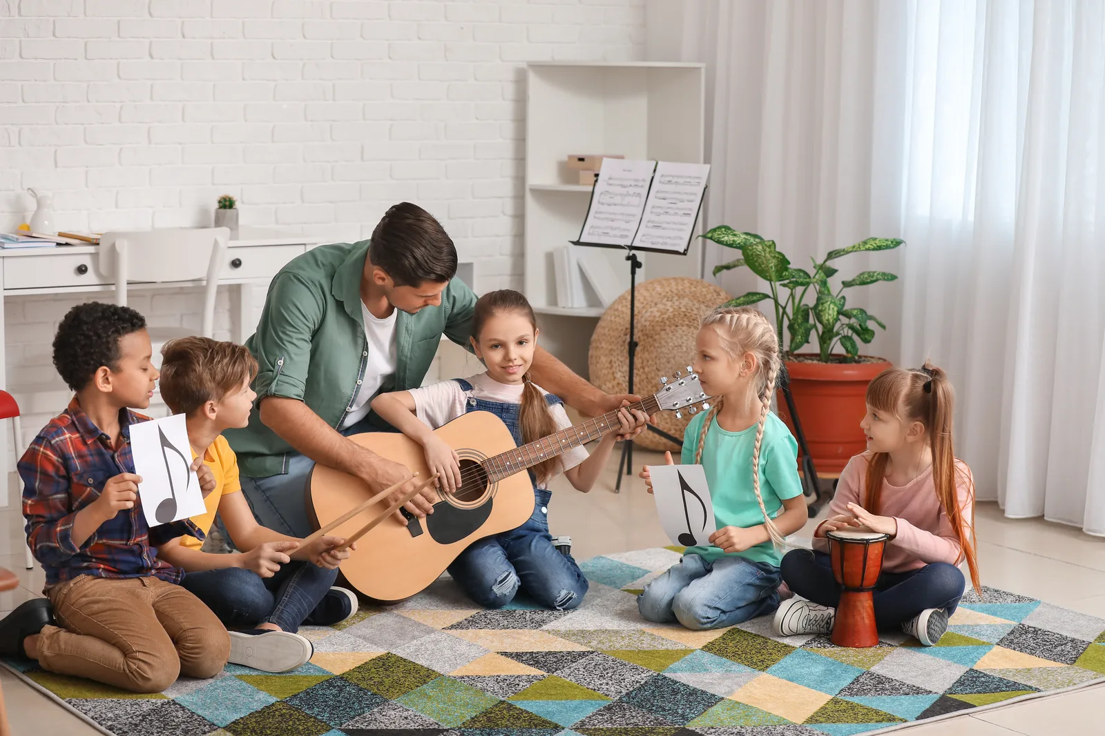 Group Music Class A music teacher helps a girl play acoustic guitar as children around them hold drumsticks, note cards and a hand drum