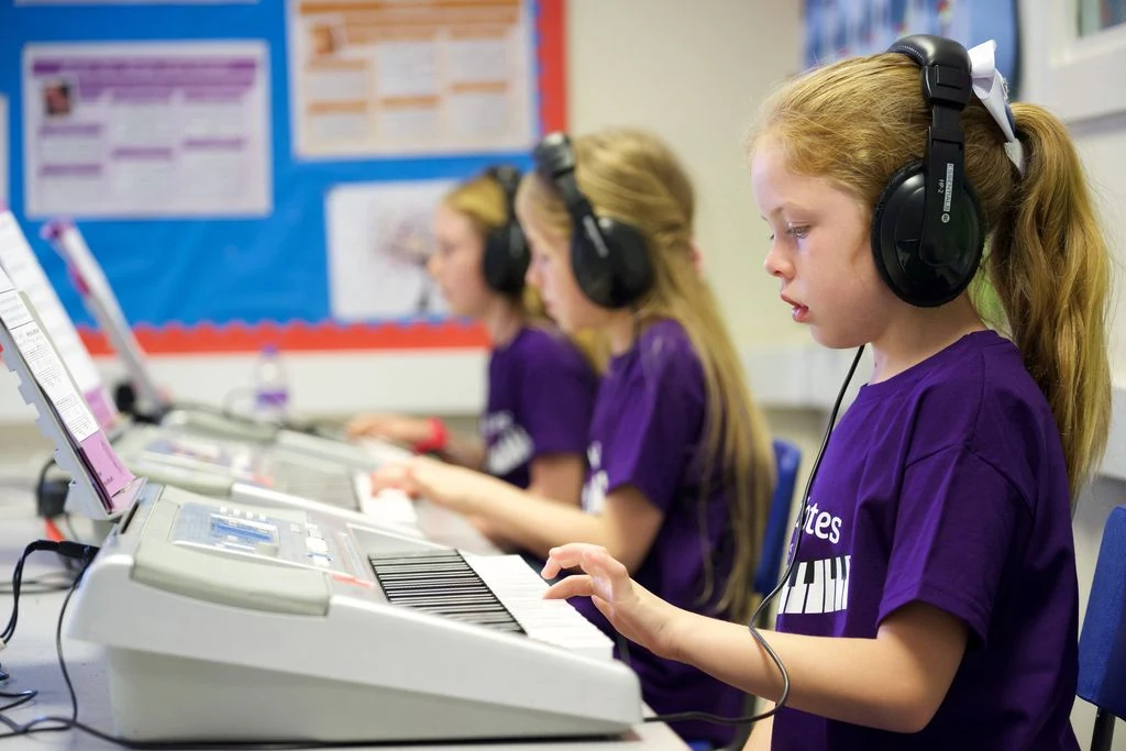 KeyNotes Students At Their Keyboards Three children in KeyNotes shirts playing electronic keyboards while wearing headphones in a group piano class