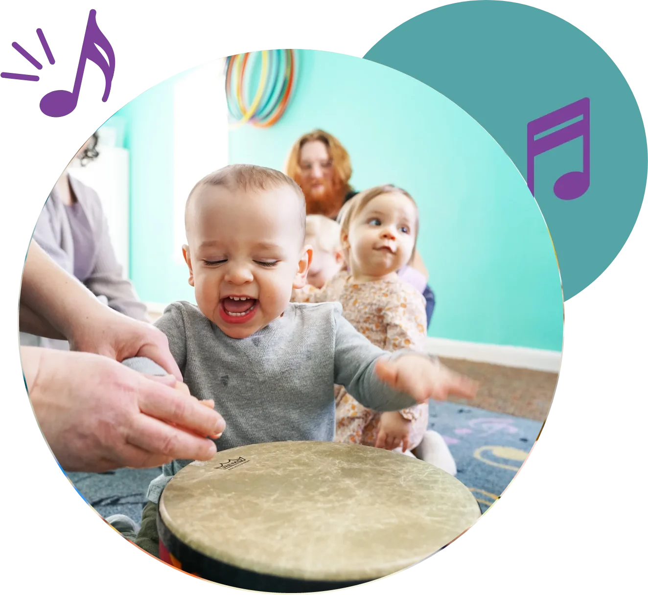 Kindermusik Baby At A Hand Drum A laughing baby pats a hand drum in a Kindermusik class as other babies and grown-ups sit on the carpet behind